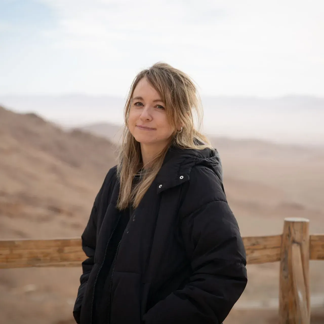 Woman in a black coat standing by a wooden fence in a desert landscape