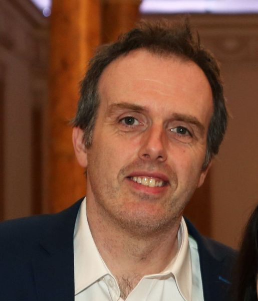Close-up of a man in a dark jacket and white shirt, smiling indoors against a wood-paneled background