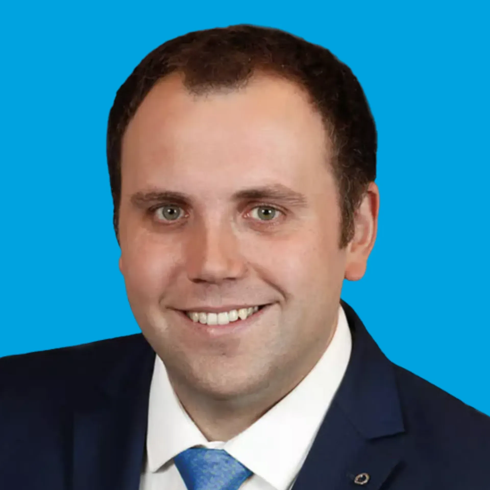 Studio portrait of a smiling man in a dark suit, white shirt, and blue tie against a blue background