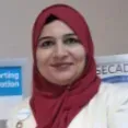 Woman in a red hijab smiling indoors, with a blue sign in the background.