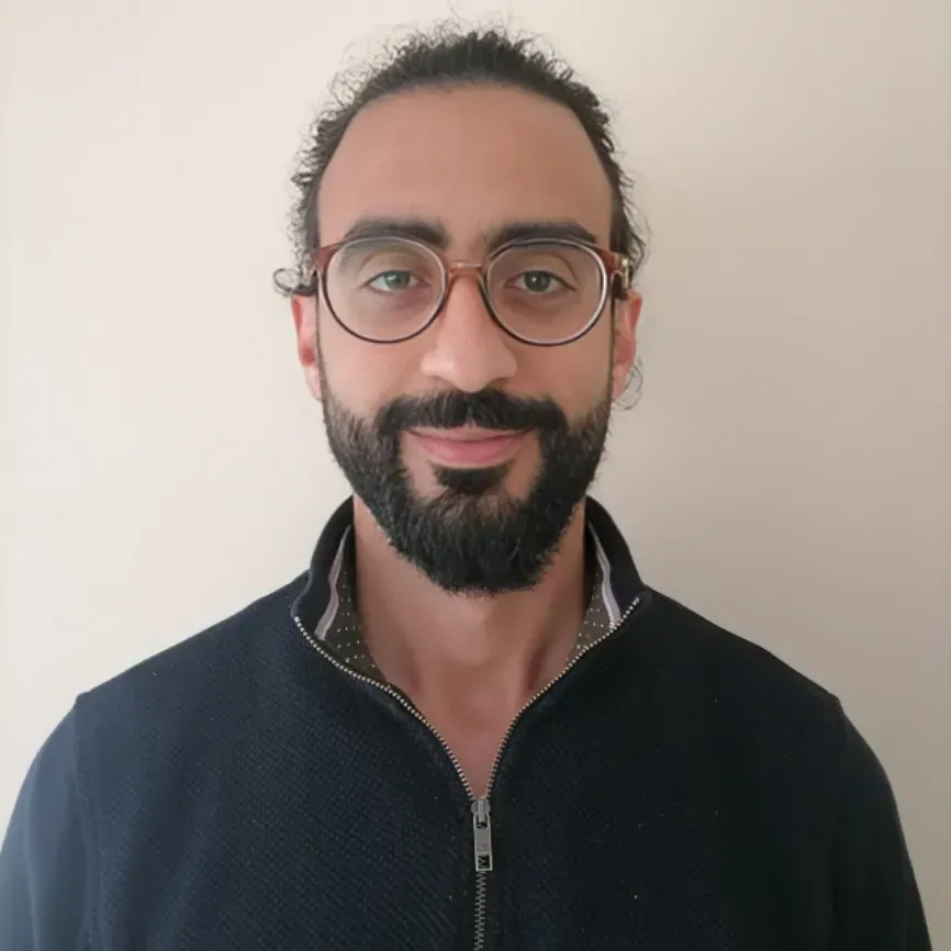 Man with glasses and a beard, wearing a dark zip-up sweater, standing against a plain wall