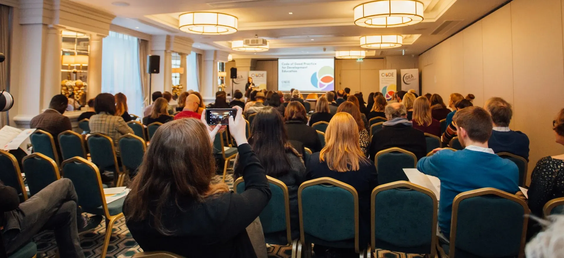 Conference audience seated in teal chairs facing a presentation screen in a hotel ballroom