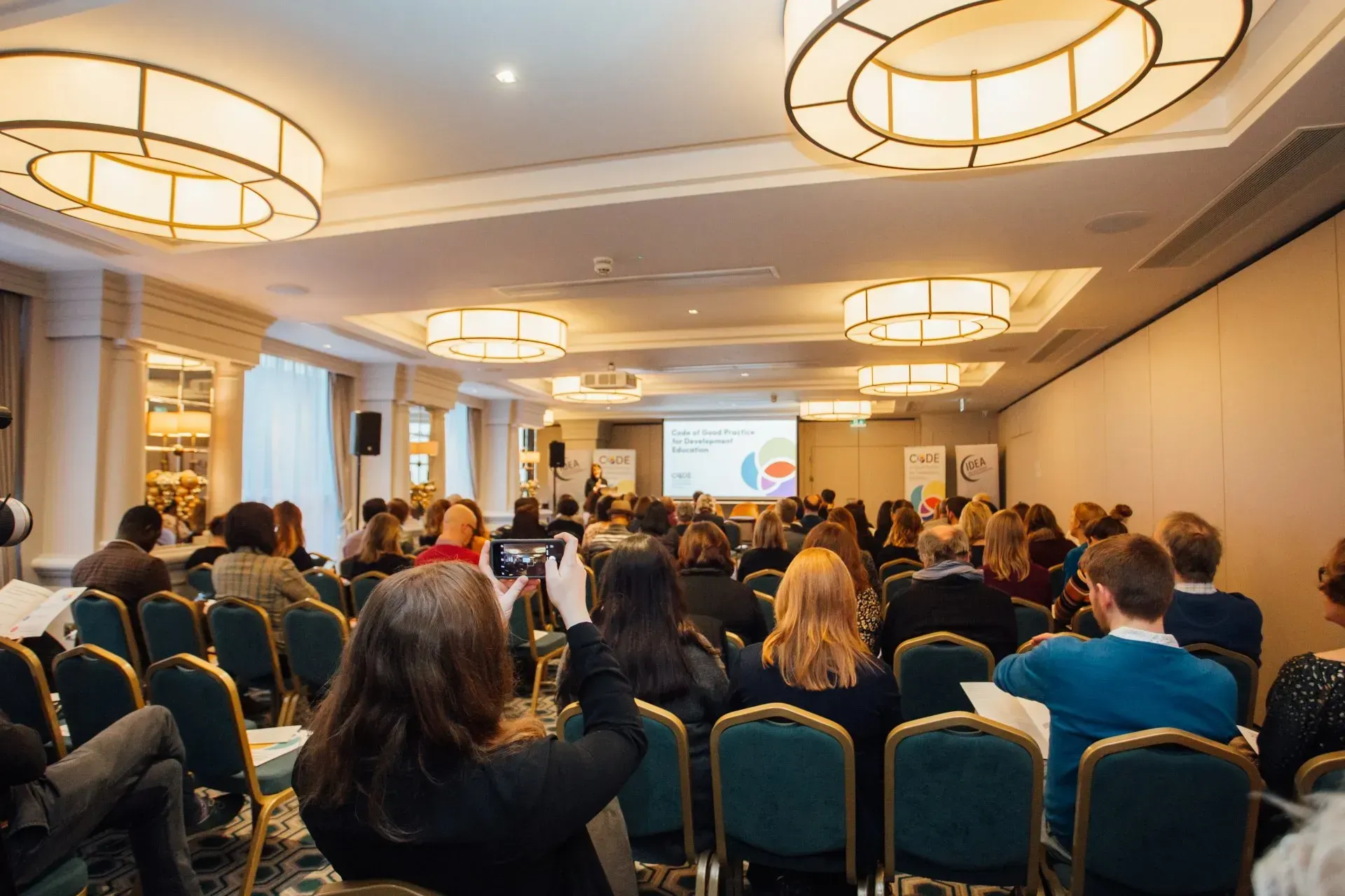 Conference audience seated in a hotel meeting room, facing a presenter at the front.