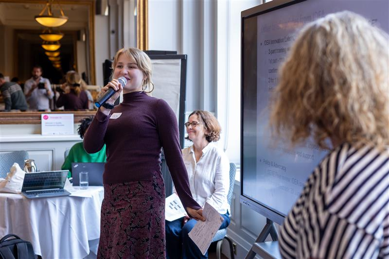 Woman speaking into a microphone at a conference, with seated attendees and a presentation screen nearby