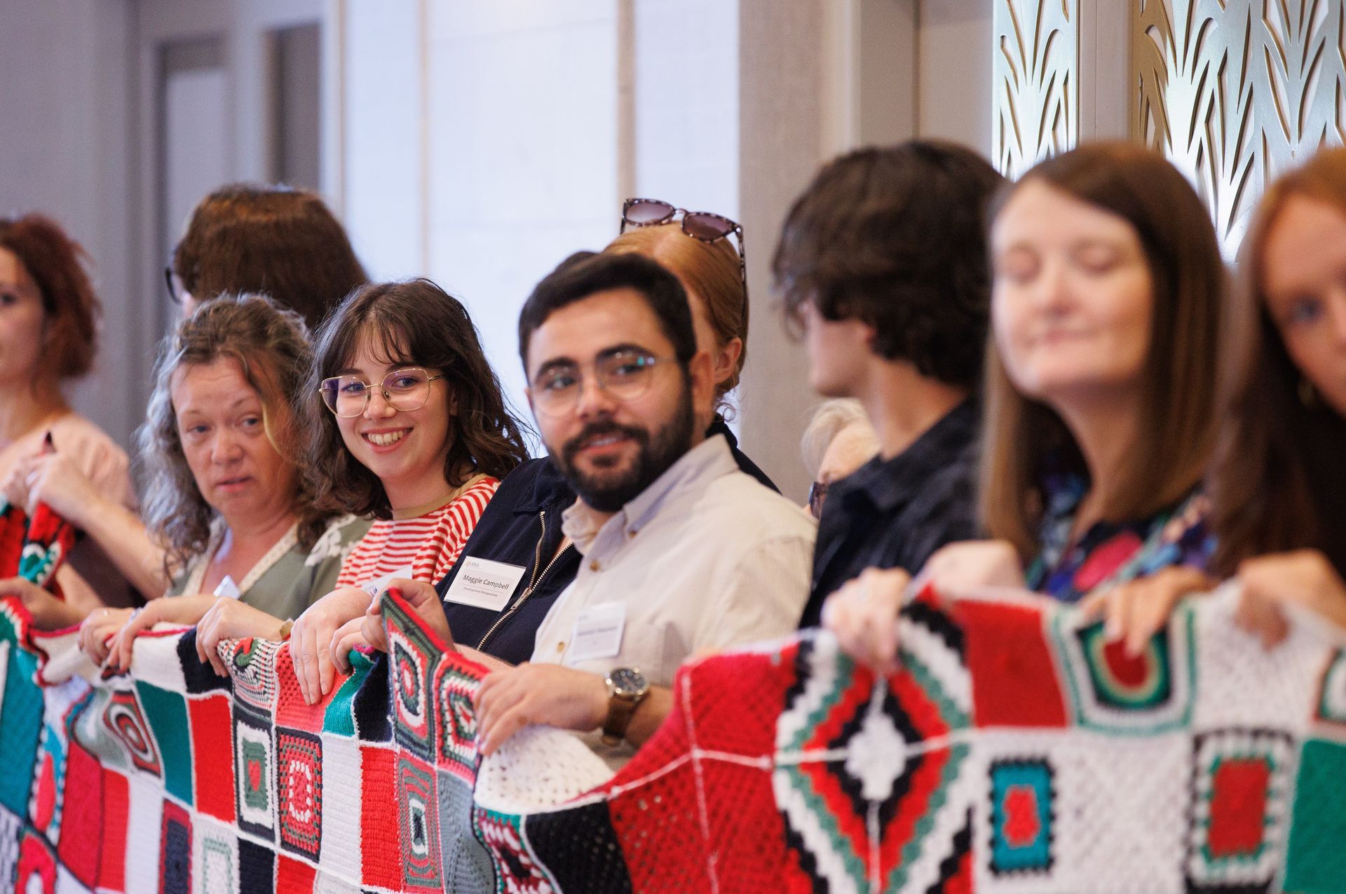 People seated behind a colorful woven banner at a community event, smiling and looking toward the camera
