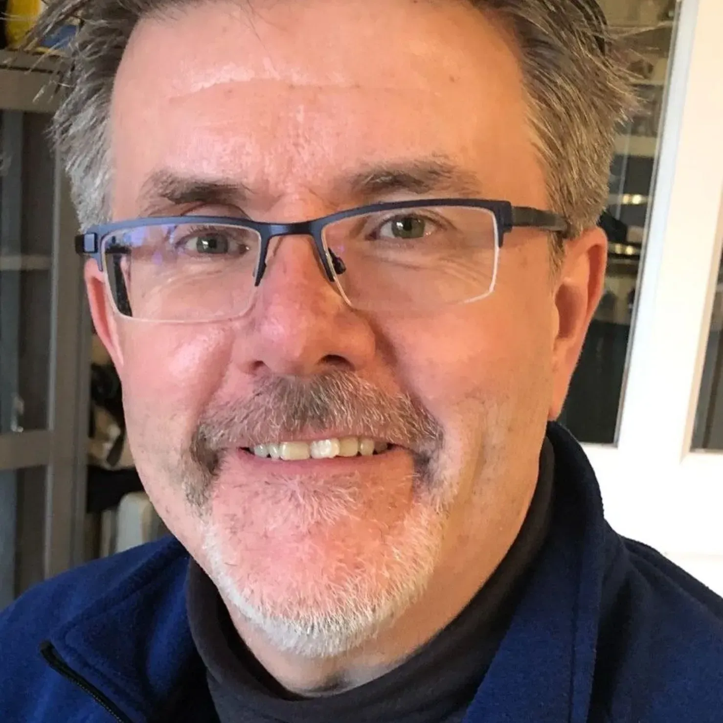 Close-up portrait of a smiling man wearing glasses and a dark shirt indoors