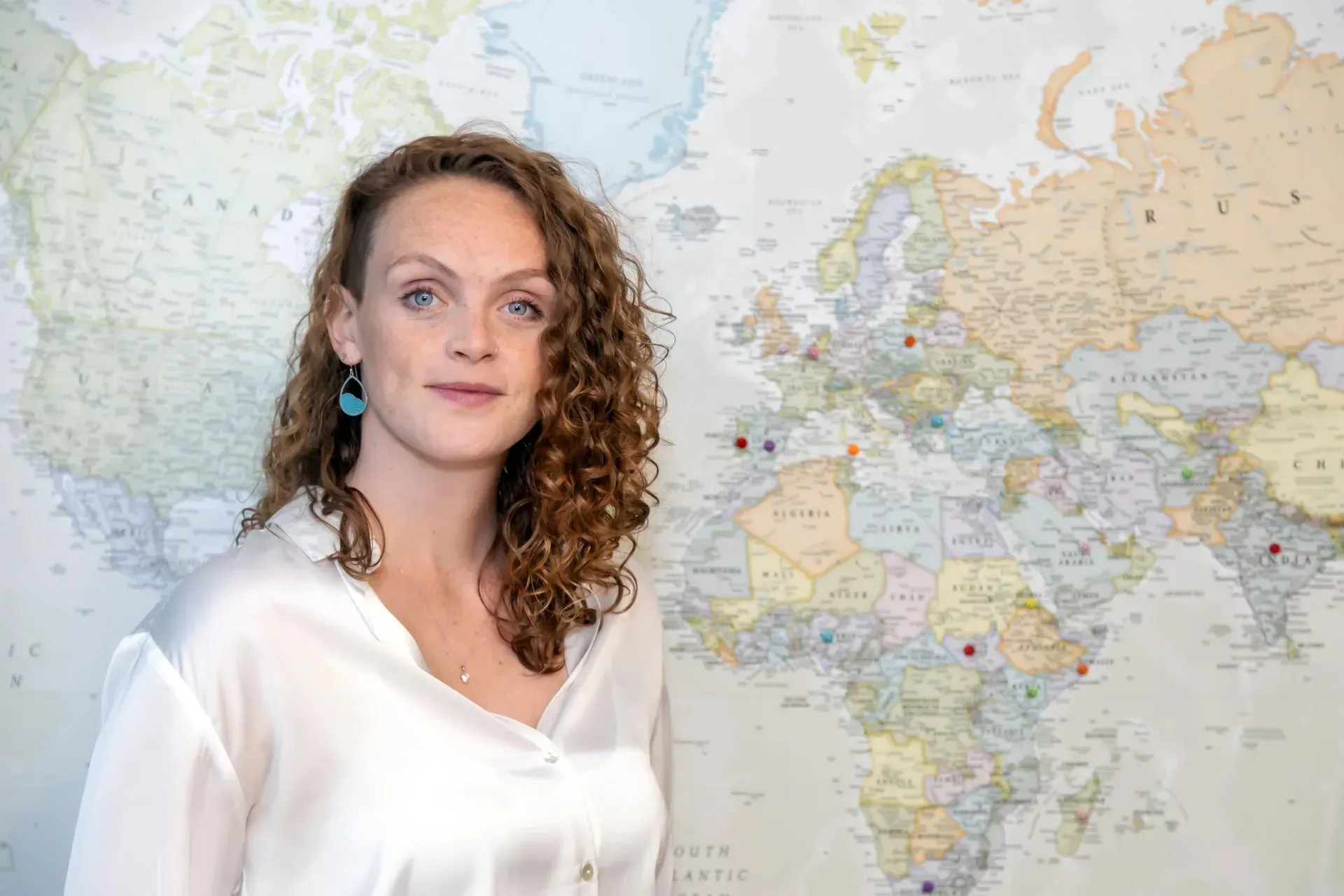 Woman in a white blouse standing before a world map, looking at the camera.