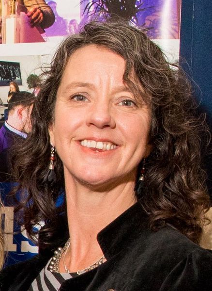 Smiling woman with curly hair at a red-carpet event, wearing a black jacket and silver necklace