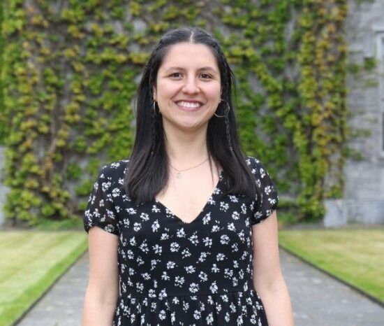 Smiling person in a black floral dress standing on a path with a green ivy wall behind.