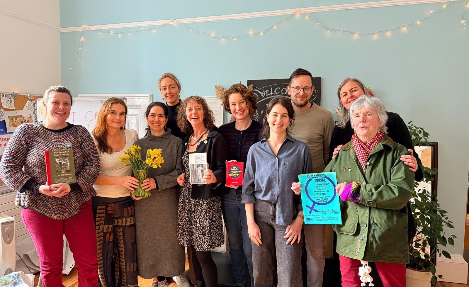Group of people standing indoors, smiling and holding flowers, candles, and colorful signs in a decorated room.
