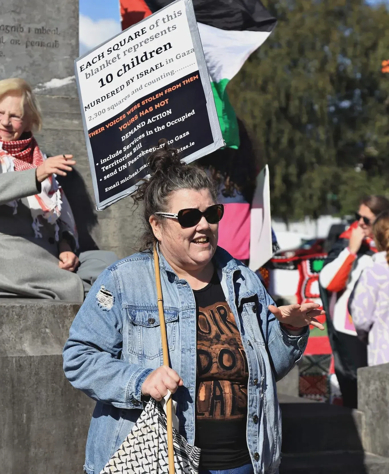 Woman in denim jacket holding a sign at an outdoor protest, with other demonstrators behind her.