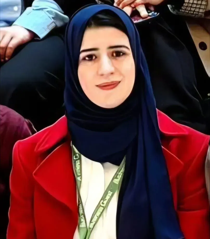 Woman in a navy hijab and red coat, smiling at an outdoor event with people blurred behind her.