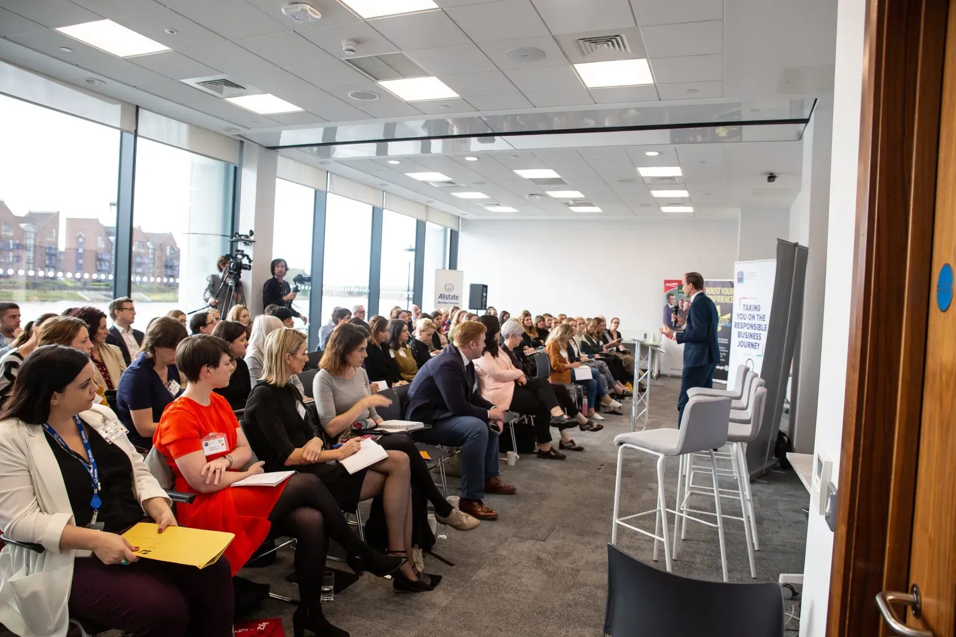 Audience seated in a bright conference room, listening to a speaker at the front.