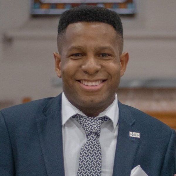 Smiling man in a navy suit and patterned tie, standing indoors.