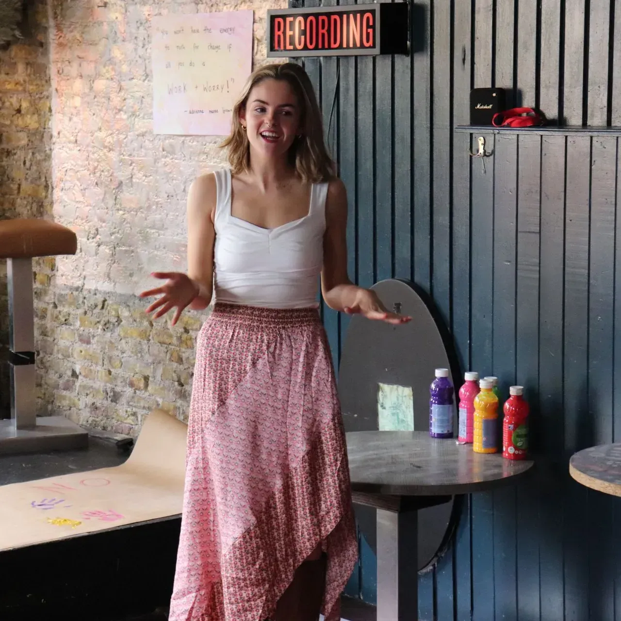 Woman standing in a recording studio, smiling beside a table with colorful bottles