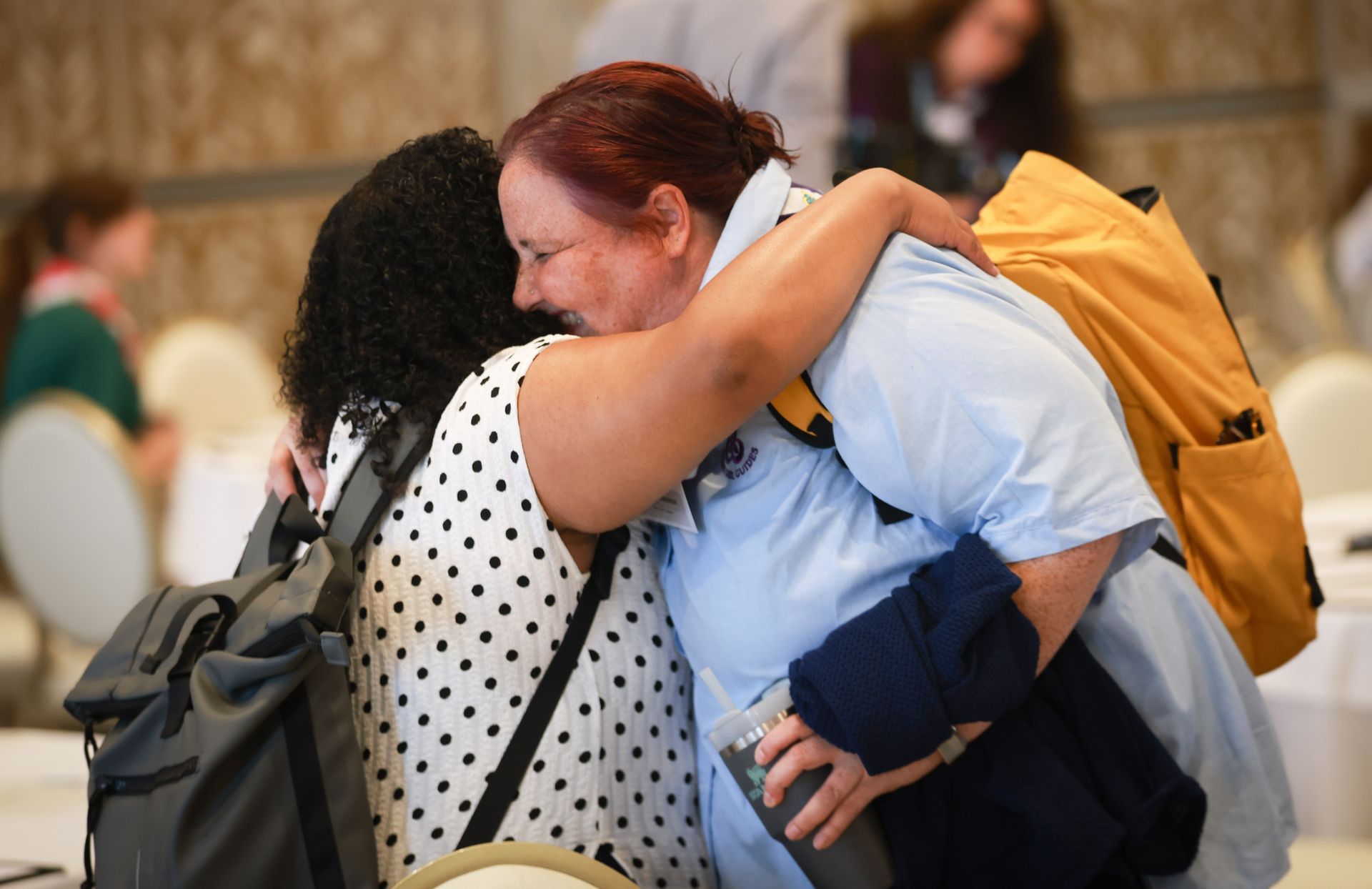 Two people hugging in a crowded indoor space, one carrying a yellow backpack