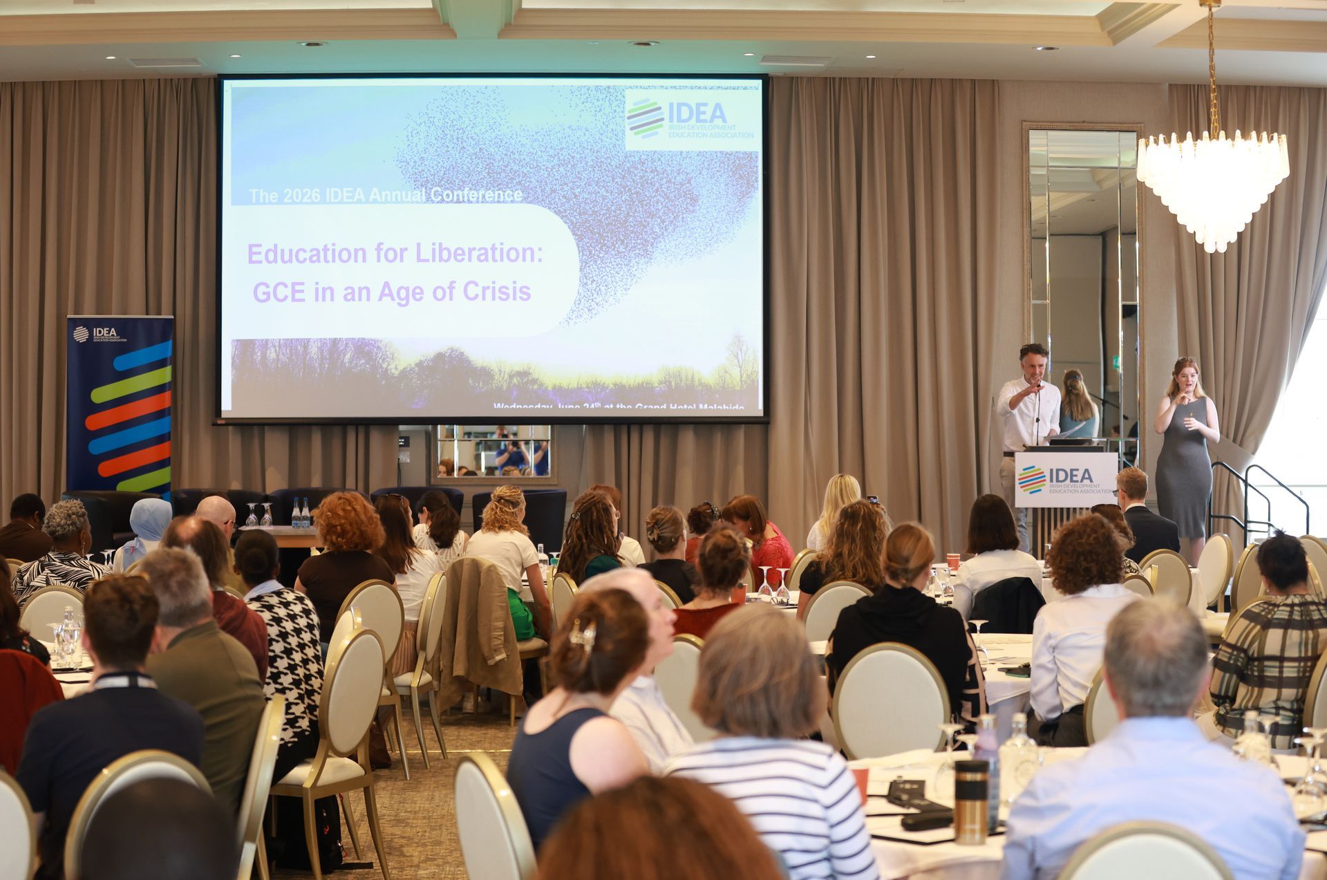 Conference audience facing a stage with speakers and a presentation screen in a banquet hall