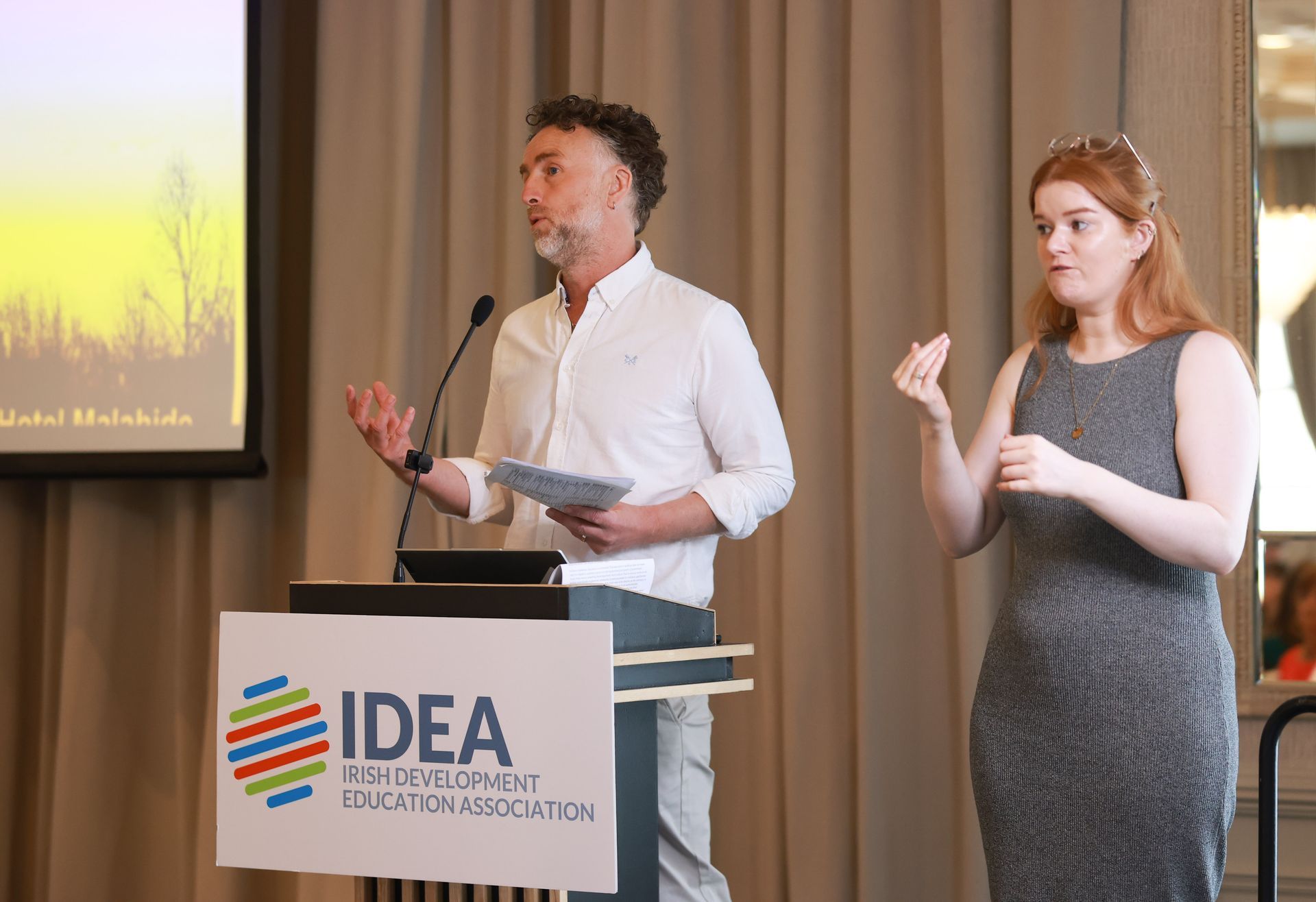 Speaker at podium with sign, presenting beside a sign language interpreter on stage.