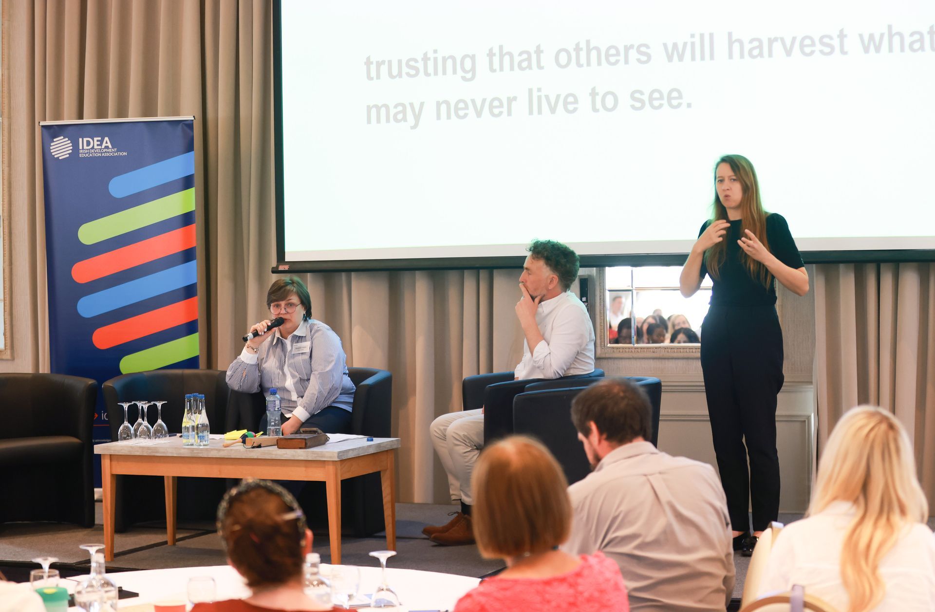 Panel discussion in a conference room, with speakers on stage and an audience listening