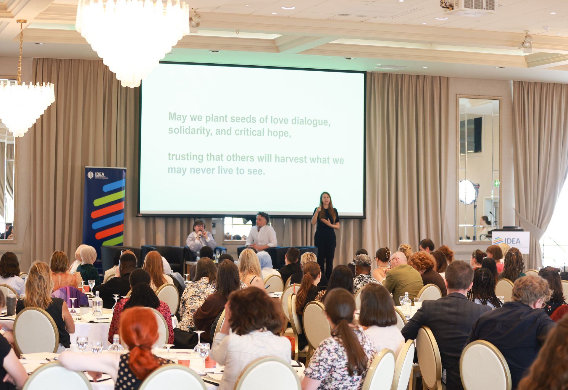 Conference audience watching a speaker on stage in a bright banquet hall with a projected presentation
