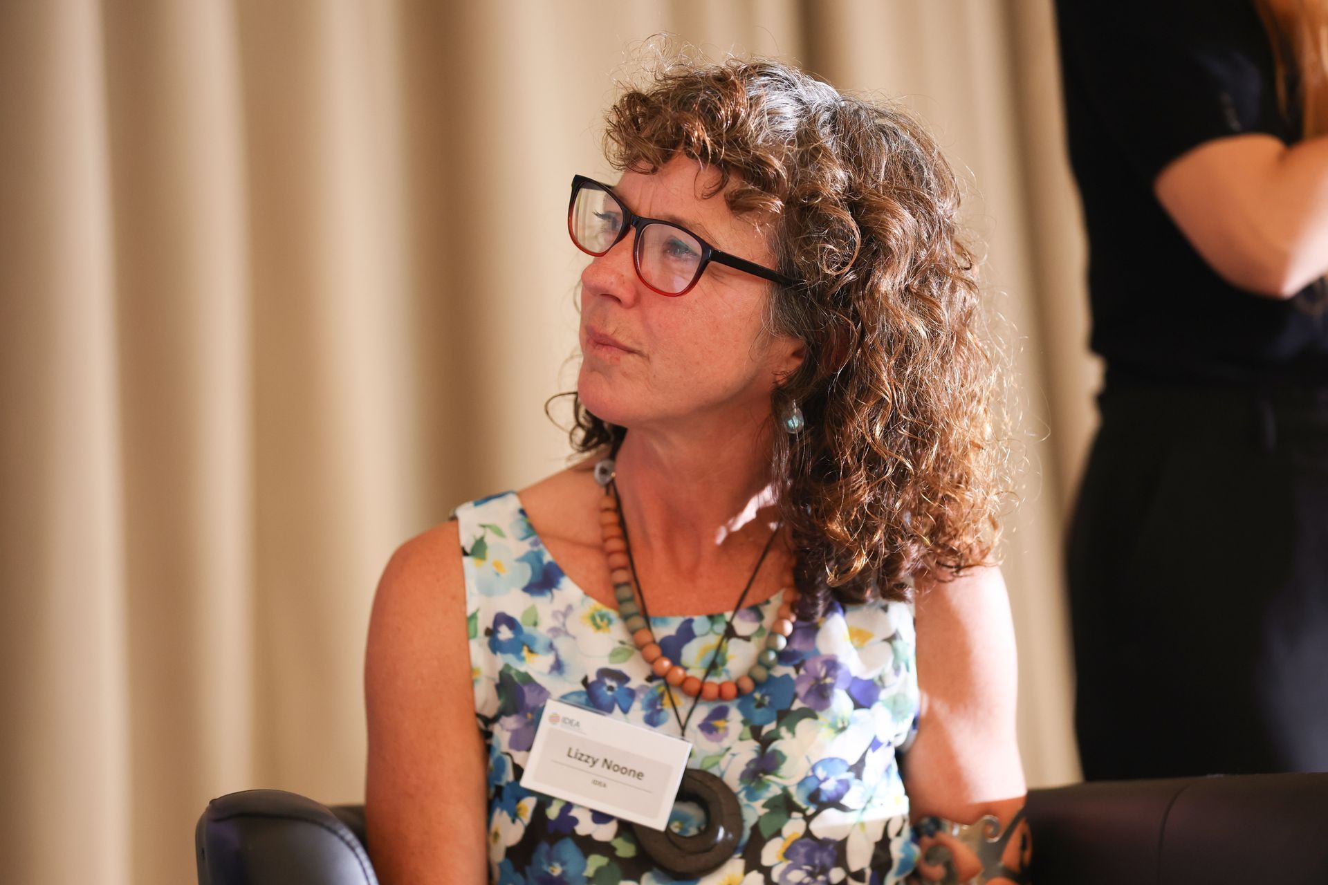 Woman with curly hair and glasses, wearing a floral dress and name badge, seated indoors and looking to the side