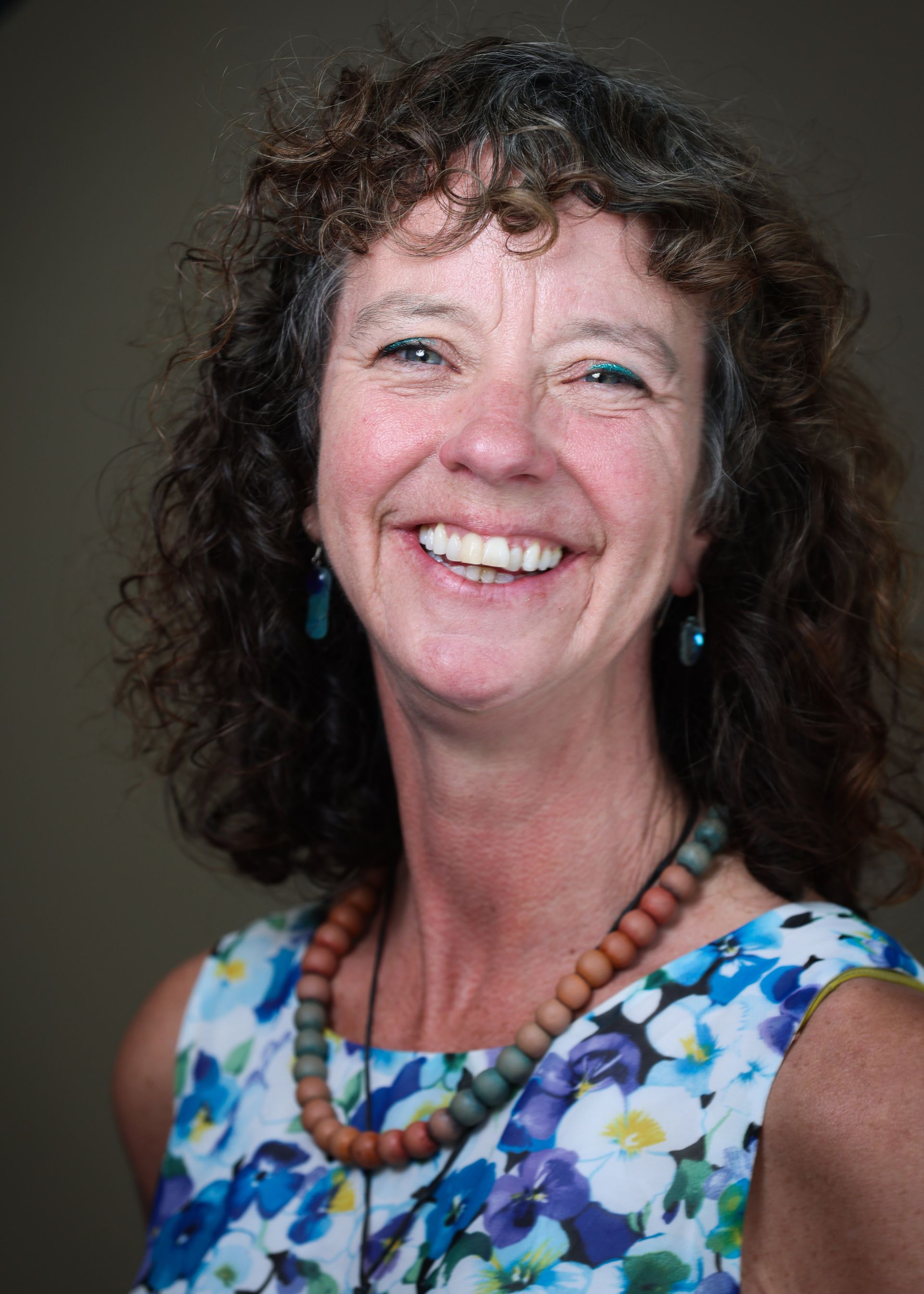 Smiling woman with curly hair wearing a blue floral top and beaded necklace against a dark background