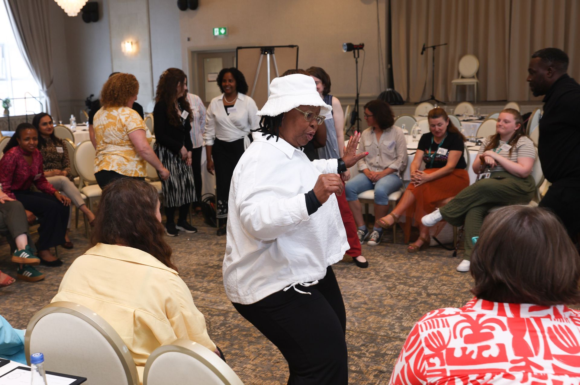 Person speaking to seated audience in a banquet hall, wearing a white hat and white shirt