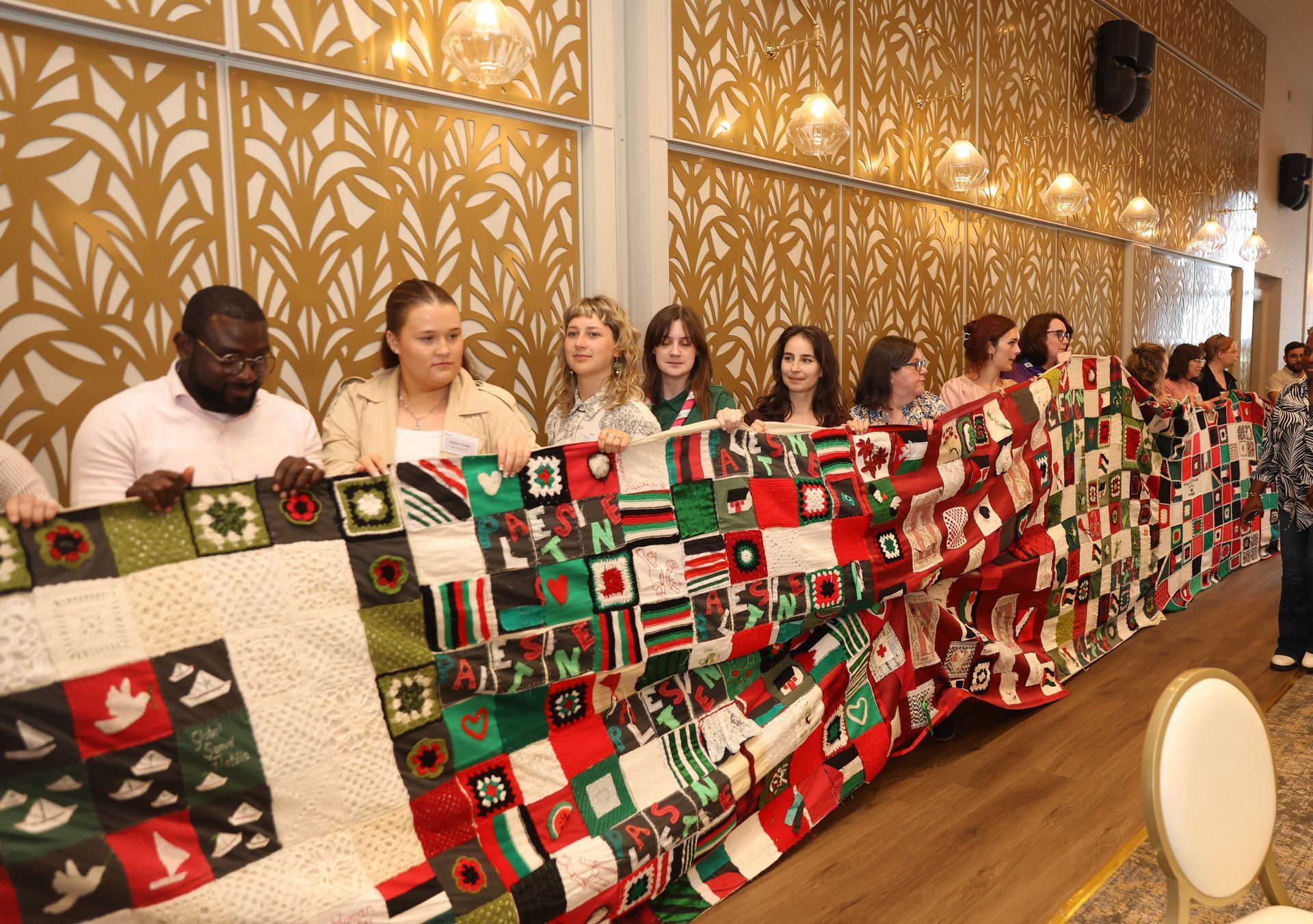 Group of people holding a long, colorful patchwork quilt in a warmly lit indoor hall