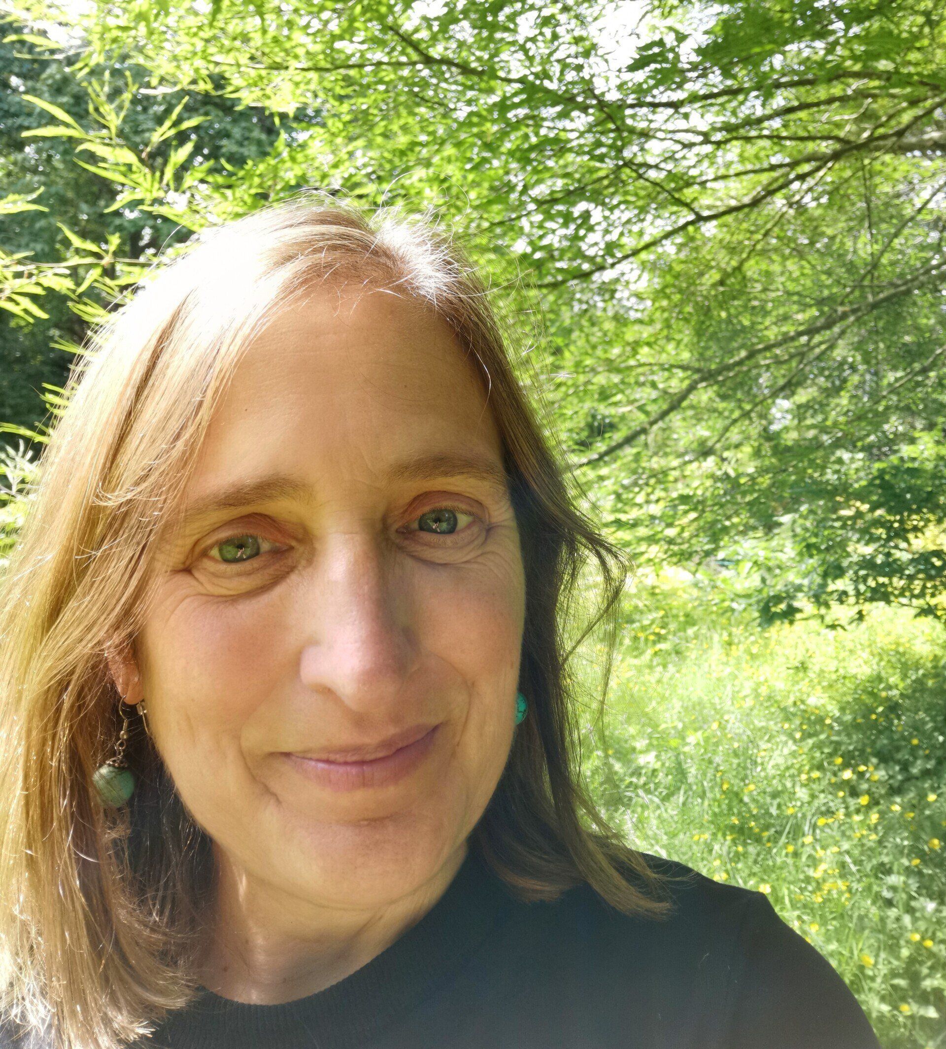 Woman smiling outdoors in a sunlit green wooded area, with trees and grass in the background