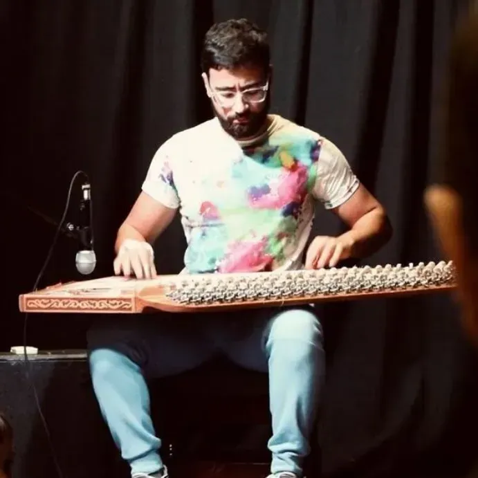 Musician seated with a qanun, playing on a dark stage, wearing a colorful shirt and glasses.