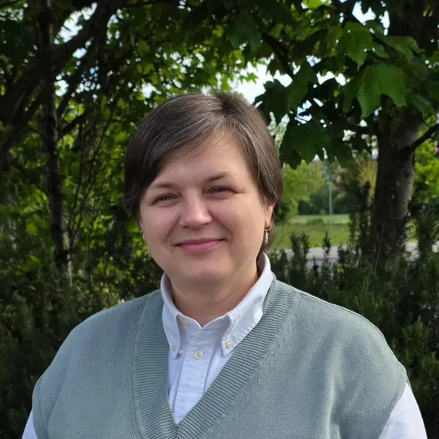 Portrait of a smiling woman outdoors in a green garden, wearing a light blue shirt and gray sweater vest