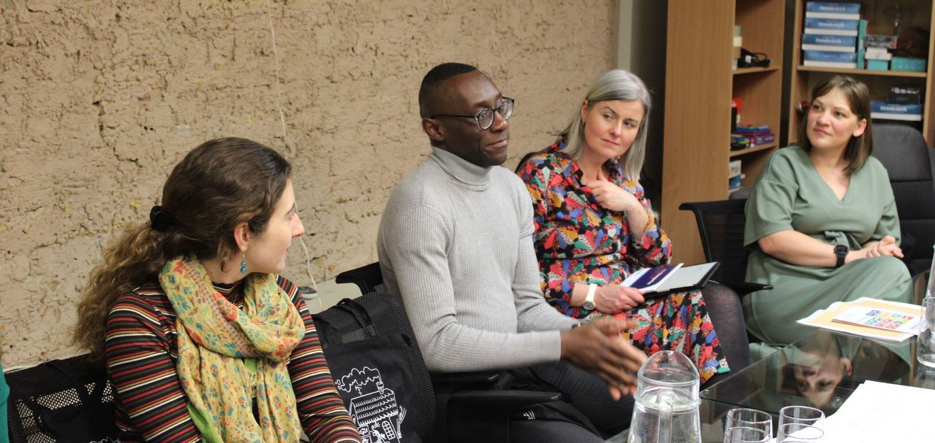 Four people seated in a discussion group, one speaking while others listen and take notes in a meeting room.