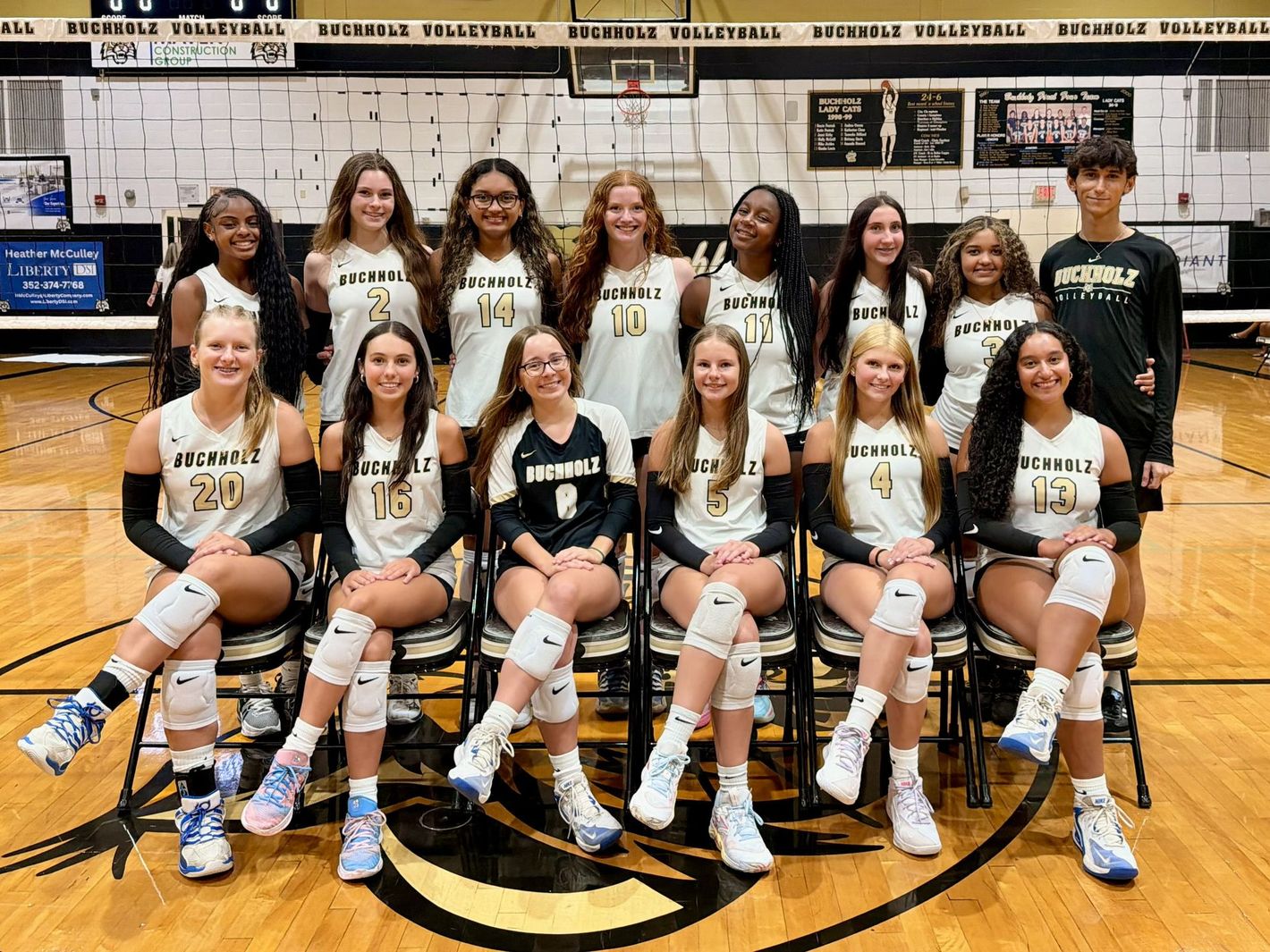 Volleyball team posing indoors in white uniforms with balls lined up at their feet.