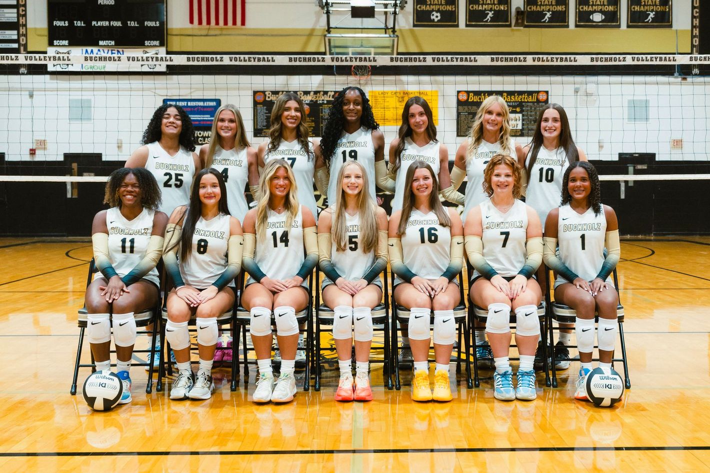 Women’s volleyball team posing in a gym, wearing white uniforms with numbers and holding volleyballs on the floor