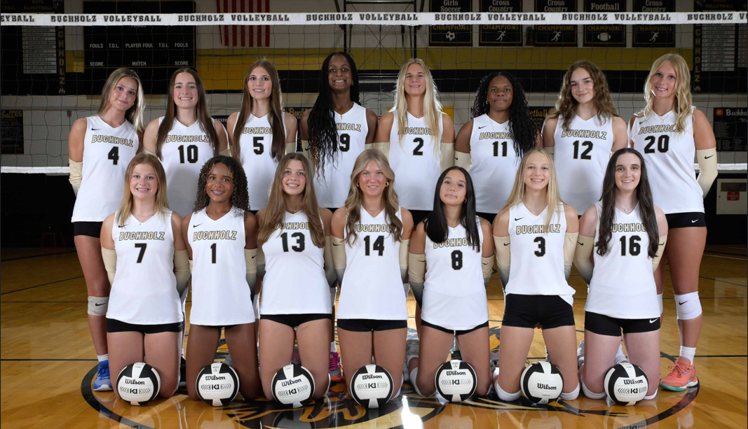Women’s volleyball team posing in a gym, wearing white uniforms with numbers and holding volleyballs on the floor