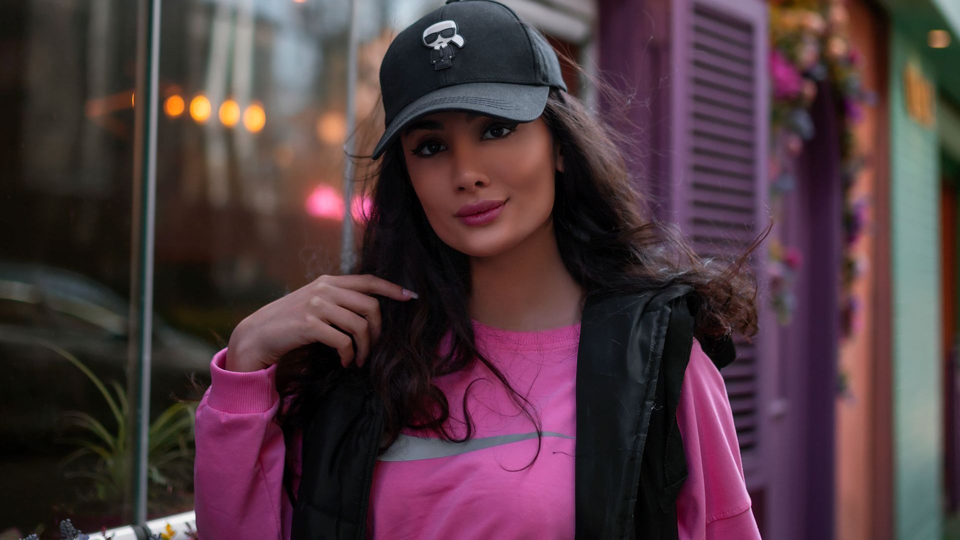A woman wearing a pink sweater and a black hat is standing in front of a purple building.