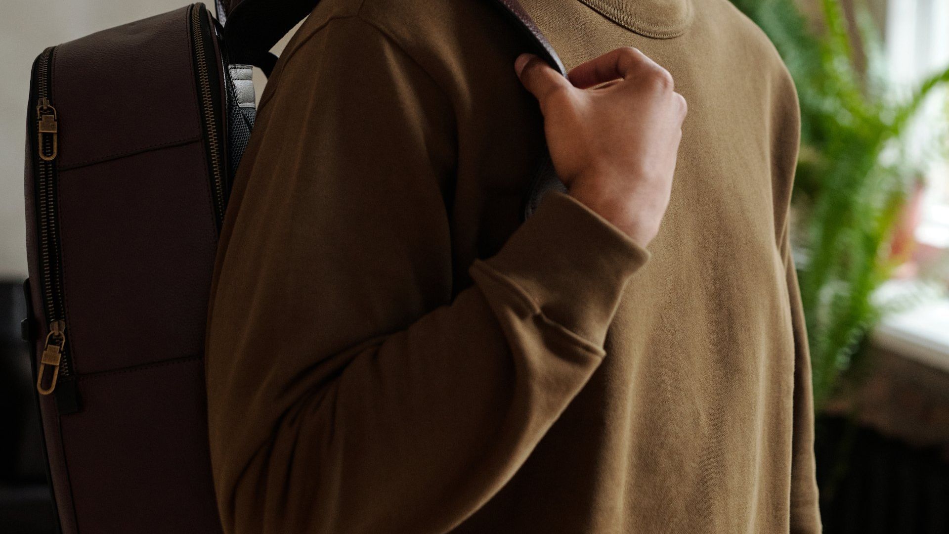 A man in a brown sweater is holding a suitcase over his shoulder.