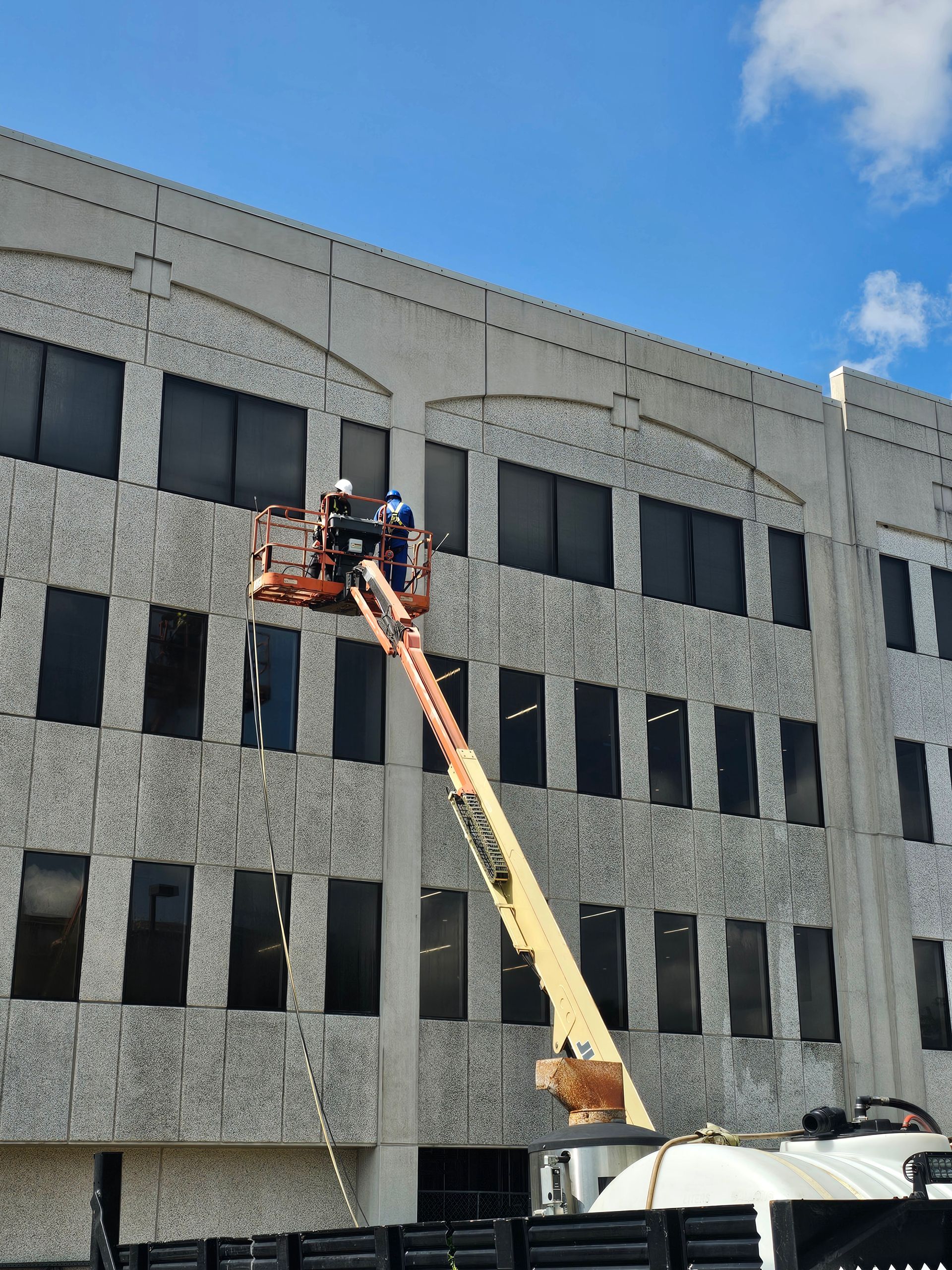 Two workers in lift platform next to a concrete building, inspecting the façade. Blue sky.