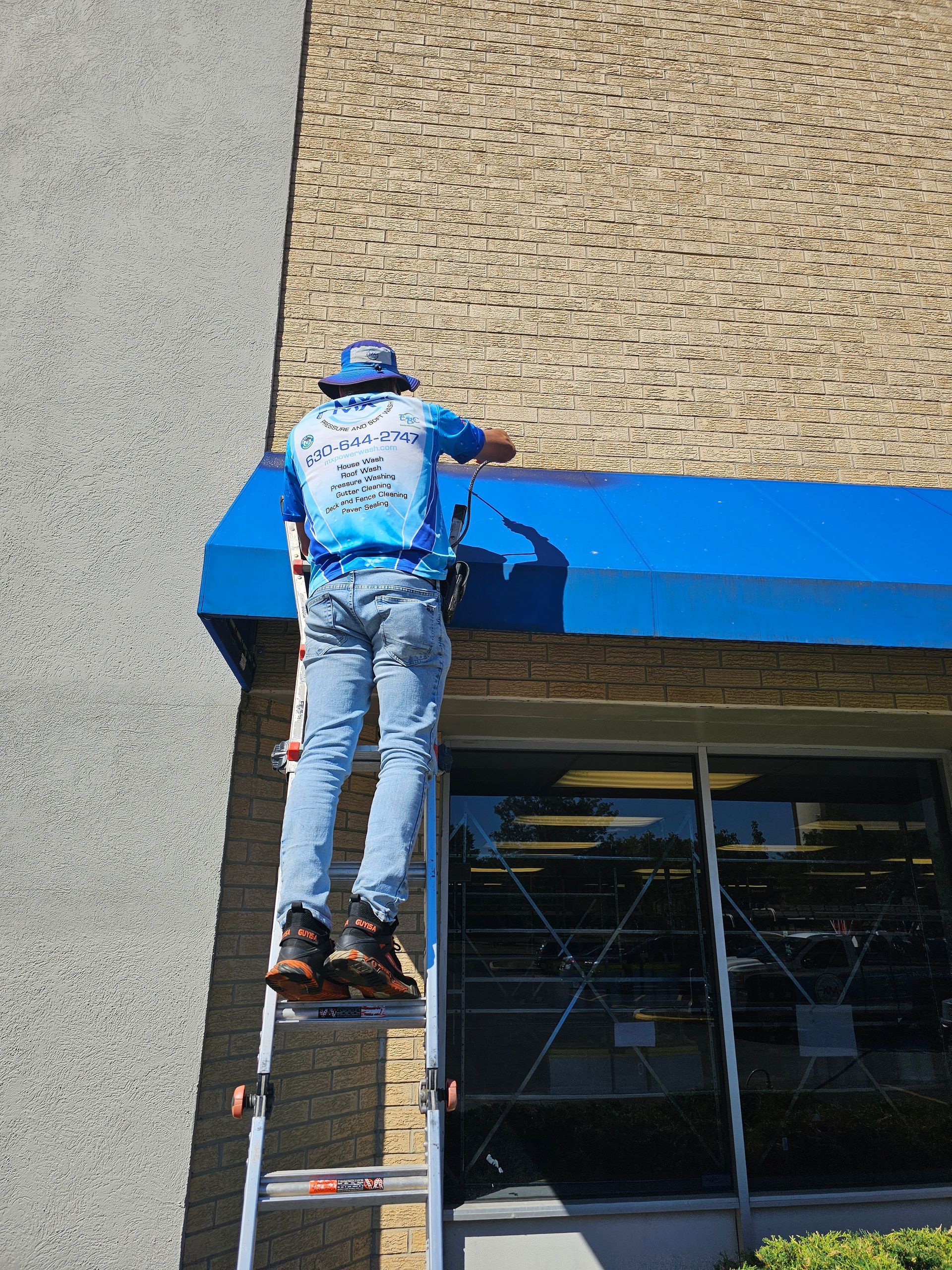 Man on a ladder repairing a blue awning. Brick building background, bright sun.