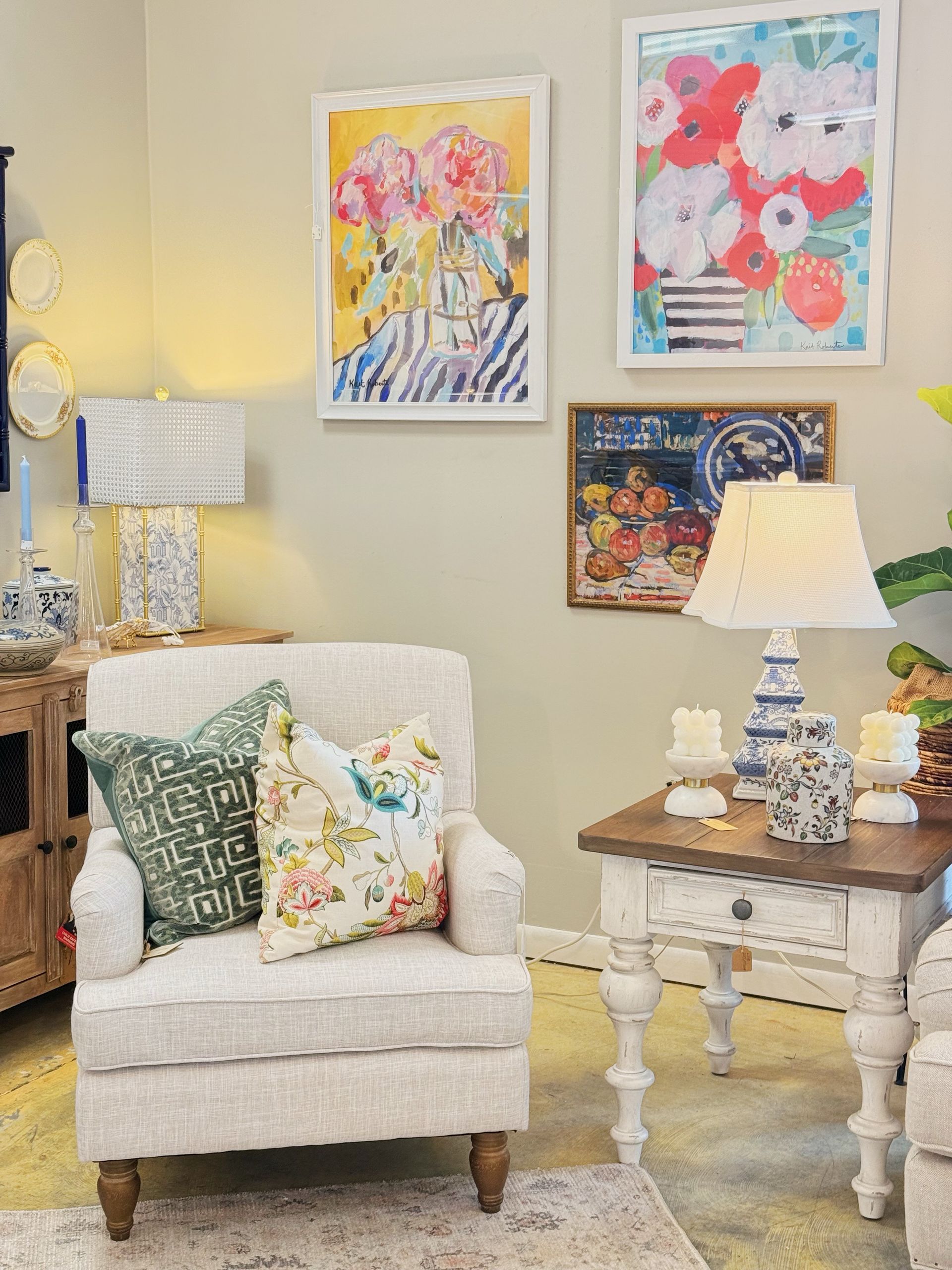 Cozy living room corner with cream armchair, patterned pillows, side table, lamps, and colorful wall art.