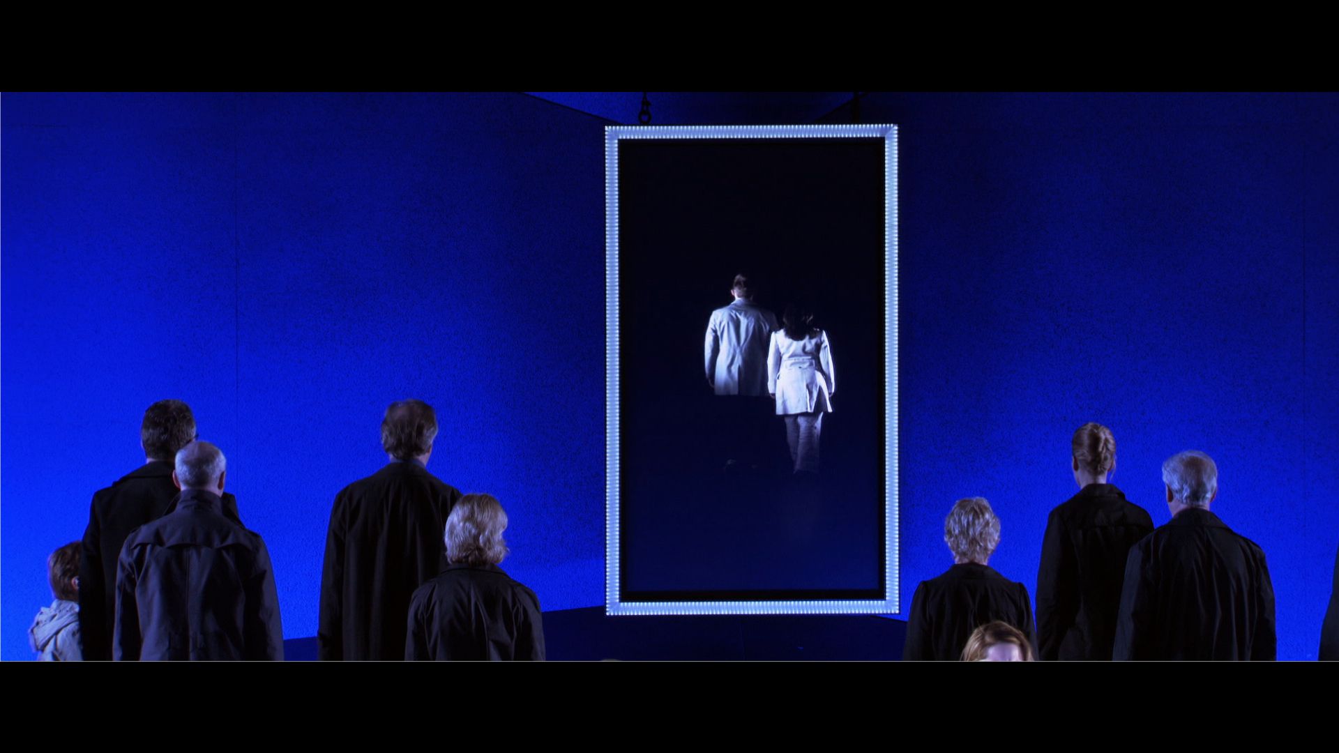 Toneeldecor met blauwe verlichting, silhouetten en twee personen in een verticaal kader.