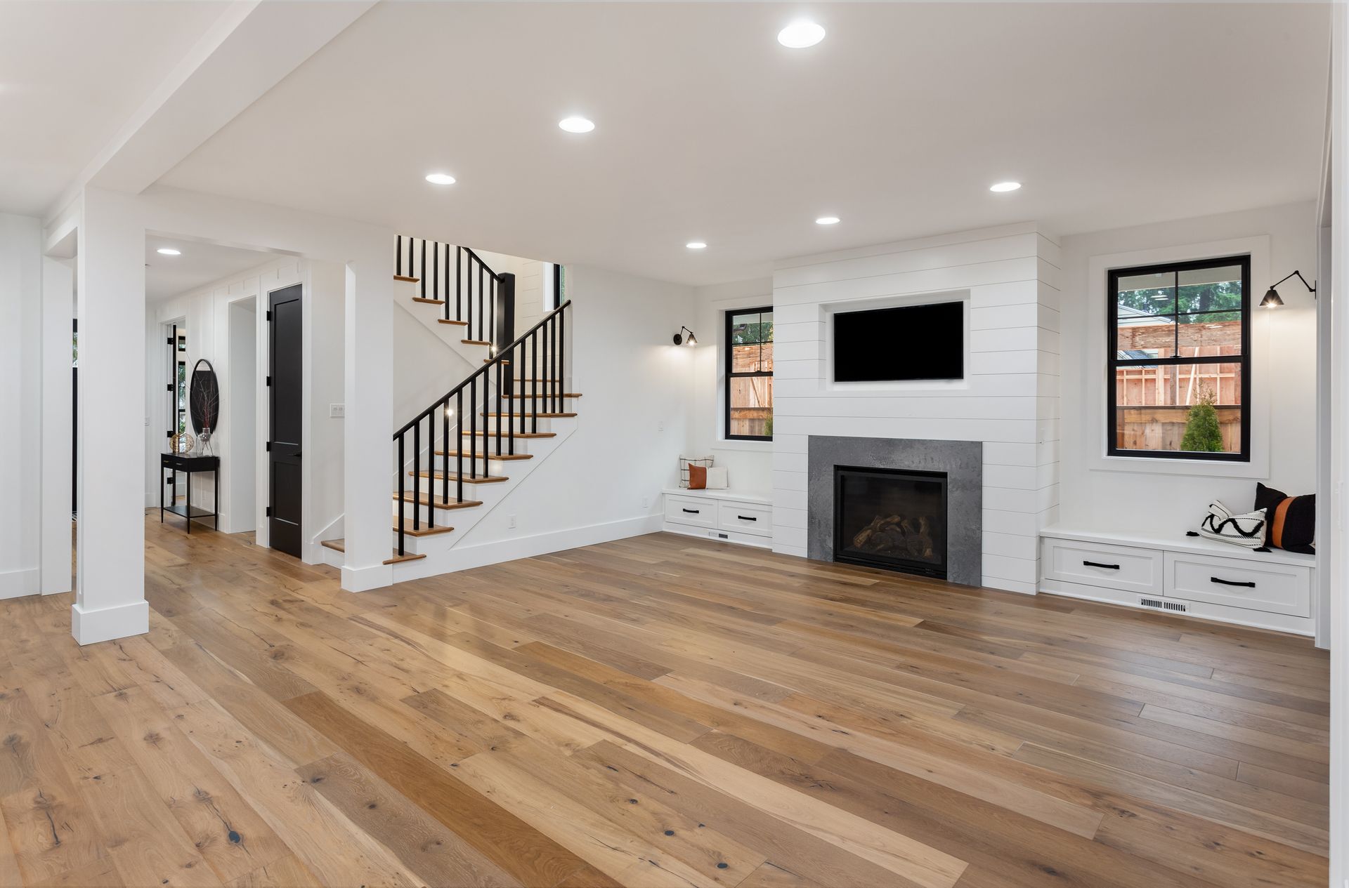 Empty modern living room with hardwood floors, fireplace, staircase, and windows.