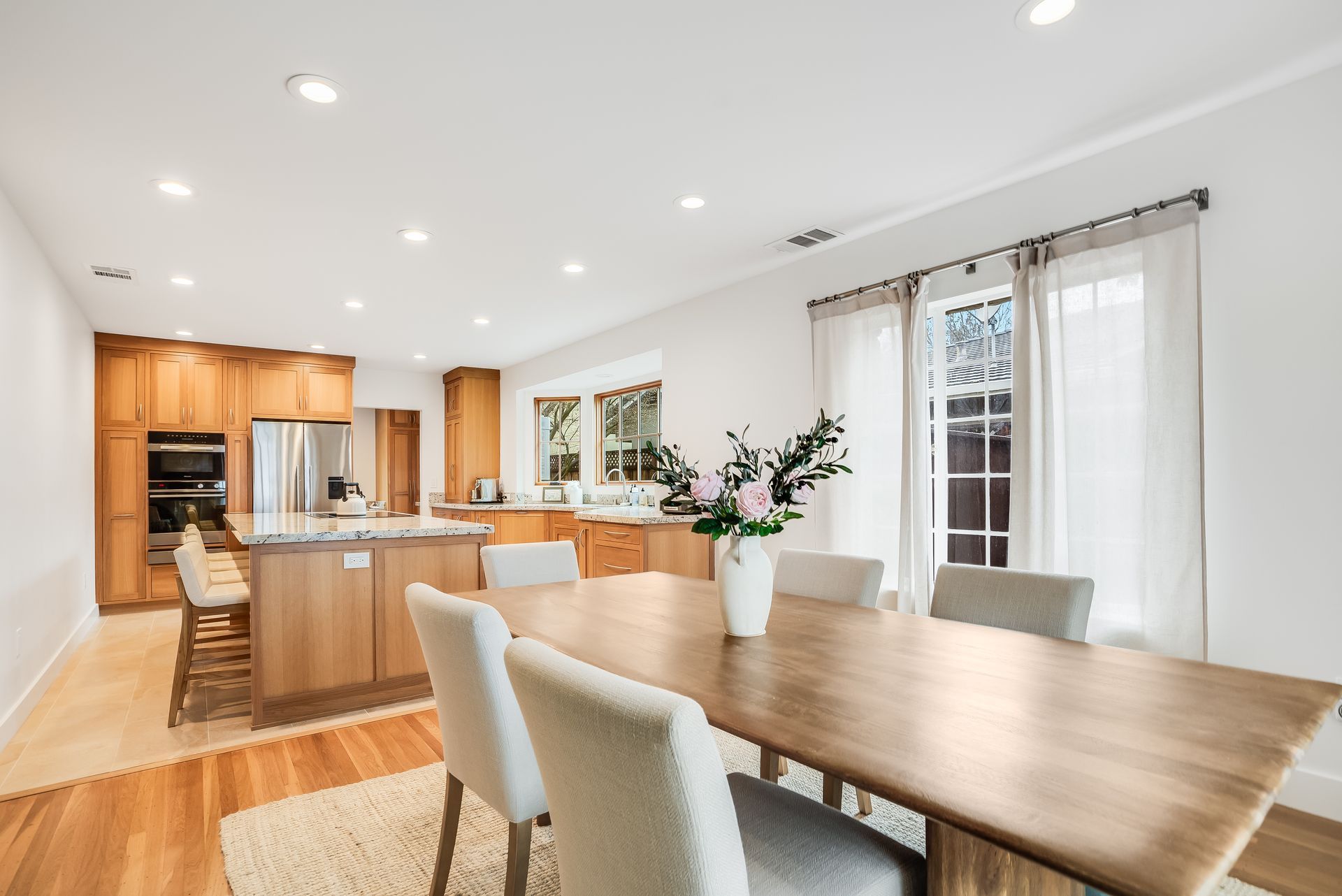 A dining room with a wooden table and chairs and a kitchen in the background.
