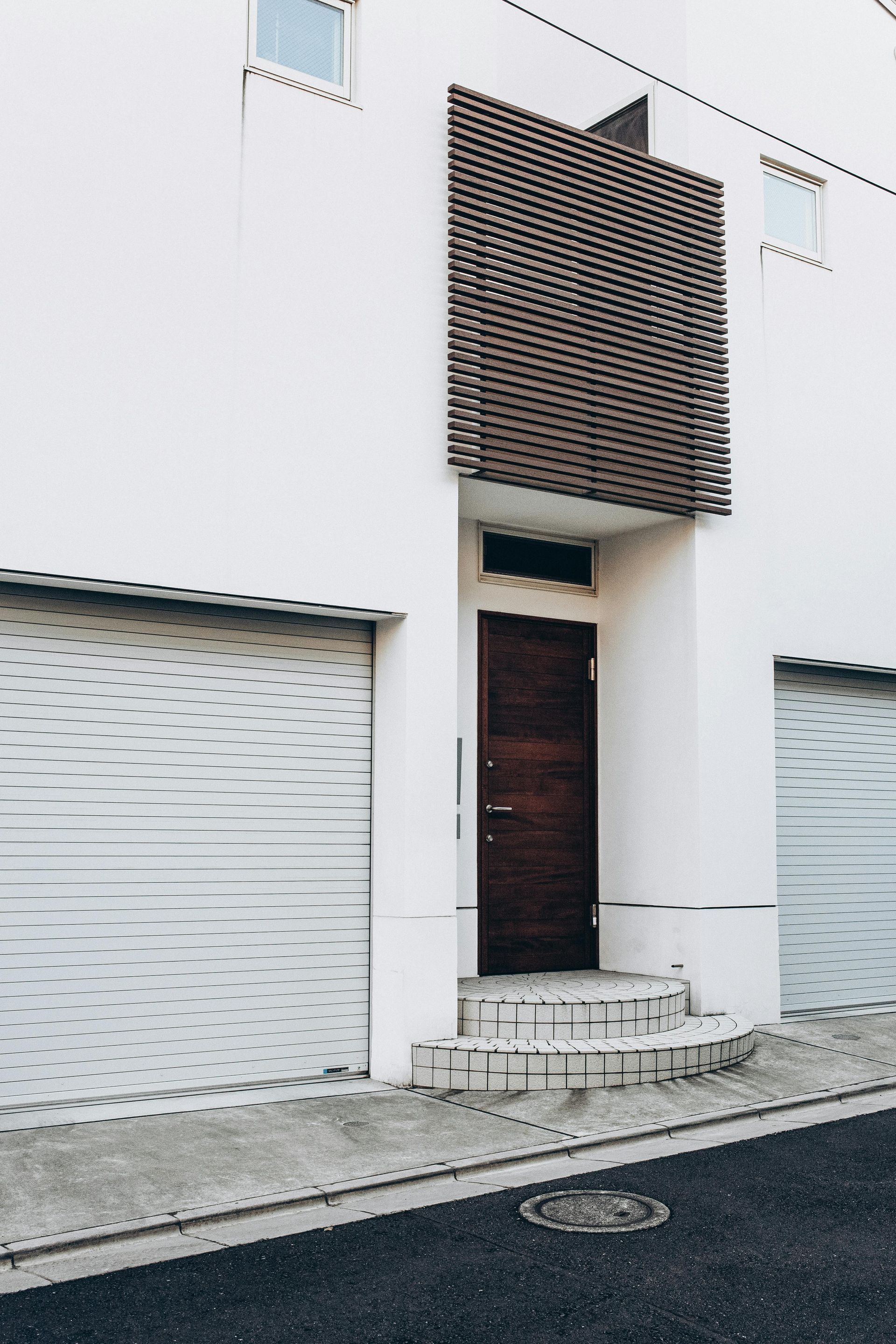 White building entrance with wooden door, trellis, and concrete steps. Two white garage doors are visible.