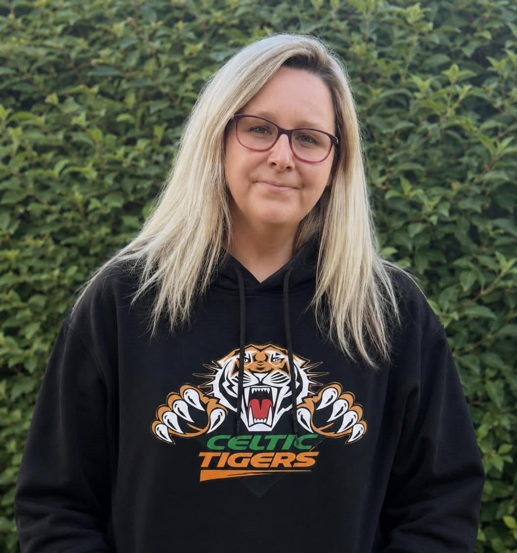 Person in a black Celtic Tigers hoodie, standing outdoors in front of green bushes.