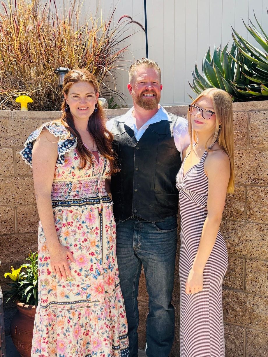 Three people standing outdoors by a stone wall, smiling in front of desert plants.
