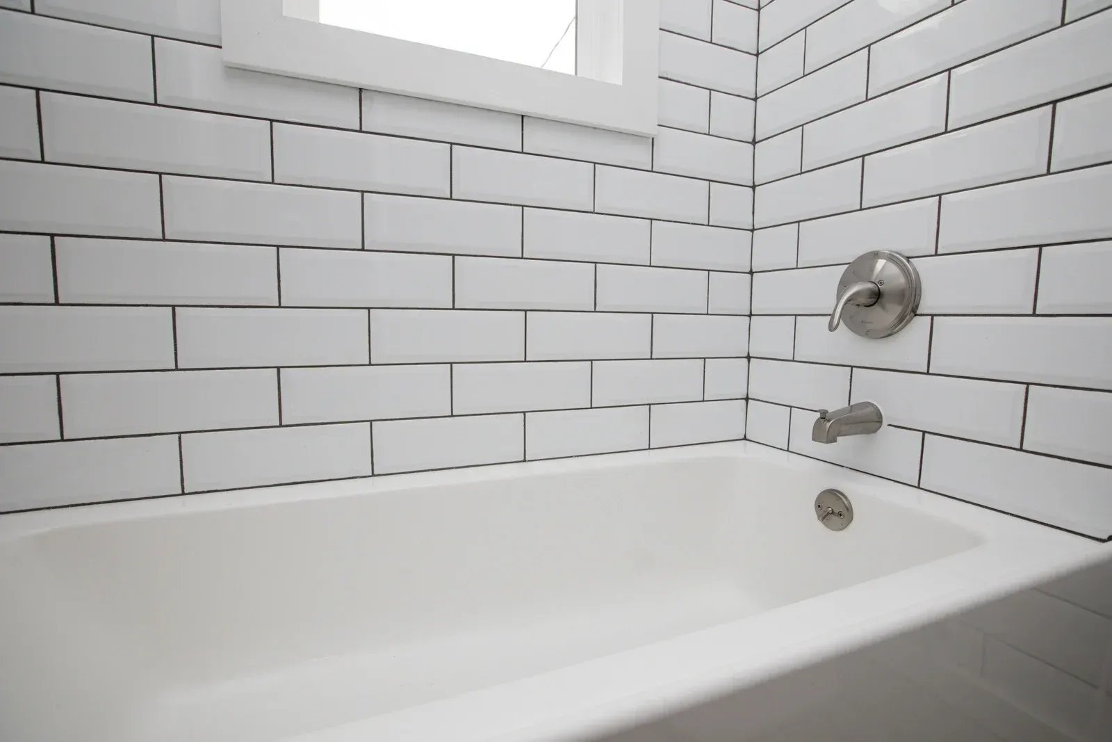 A bathtub in a bathroom with white tiles and a window.