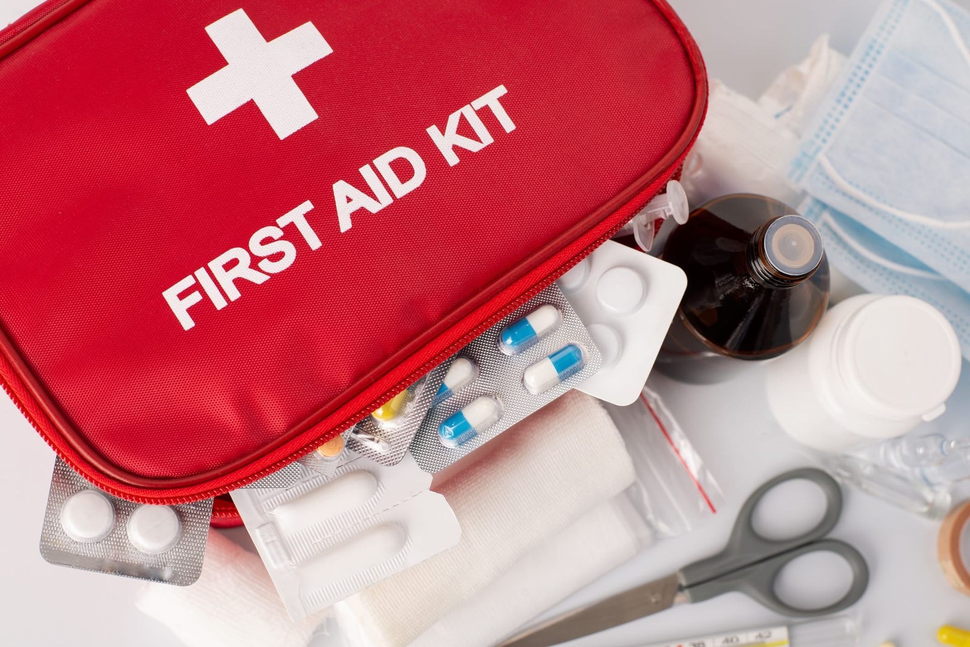 Red first aid kit with medical supplies, pills, bandages, scissors, and medicine bottles on a white surface