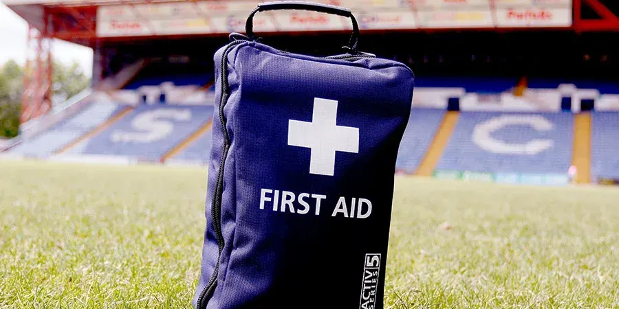 Blue first aid kit with white cross and “FIRST AID” text on a sports field sidelines.