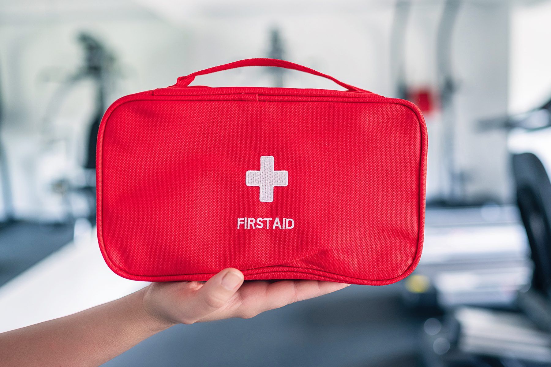 Red first aid kit with white cross held in hand, against a blurred gym background